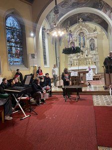In einer Kirche sitzen eine Gruppe von Menschen vor einem Altar, während andere hinter einem Klavier und einem Podium stehen. Ein Priester spricht, und eine Kronleuchter hängt von der Decke.