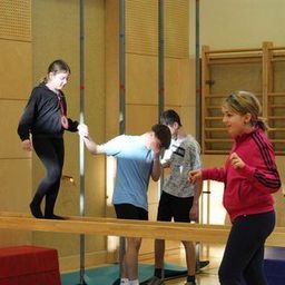Vier Kinder, zwei Mädchen und zwei Jungen, üben Balance auf einer schmalen Stange in einer Turnhalle.