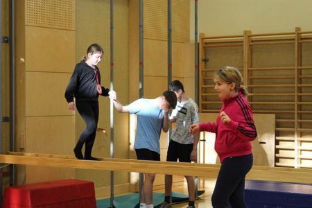 Vier Kinder, zwei Mädchen und zwei Jungen, üben Balance auf einer schmalen Stange in einer Turnhalle.