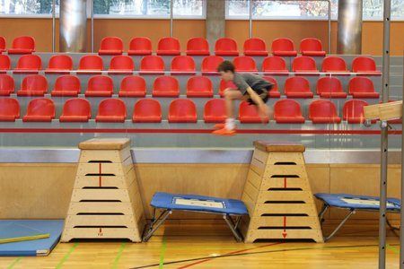 Ein Junge springt in einer Turnhalle über Hürden, mit roten Stühlen im Hintergrund.