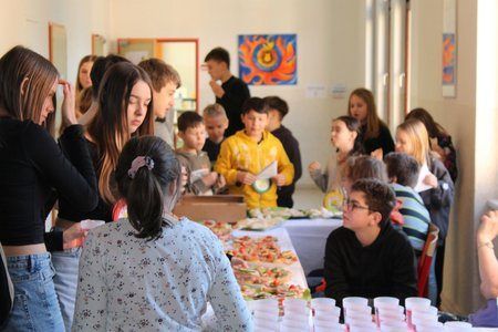 Mehrere Kinder und Erwachsene stehen in einem Raum, scheinbar auf einer Party. Einige Kinder stehen um einen langen Tisch mit Essen. Ein älterer Junge sitzt am Tisch.