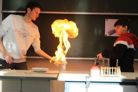 Zwei Personen befinden sich in einer Laborumgebung, eine hält eine Schale mit Flammen, während die andere mit einem Becher hilft.