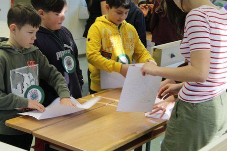 Eine Gruppe von Kindern in einem Klassenzimmer, die verschiedene Jacken tragen, arbeiten an einem Projekt mit Papier und Stiften auf einem Holzschreibtisch. Eine Frau unterstützt sie.