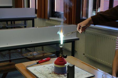 Eine Person benutzt einen Bunsenbrenner in einem Klassenzimmerlabor mit einer brennenden Flamme darüber. Die Einrichtung umfasst einen Holztisch, ein Glasbecher und einen roten Schraubendreher.