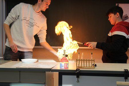 Zwei Schüler arbeiten an einem Chemieexperiment mit einer Flamme. Ein Junge mit einem schwarzen und roten Jacke hält einen Stock, während ein anderer Junge in einem weißen Shirt beobachtet.