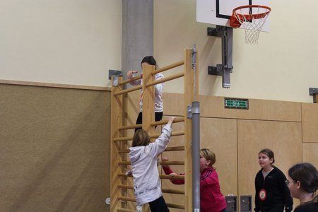 Drei Mädchen klettern in einer Turnhalle auf eine Holzleiter. Zwei weitere Mädchen stehen daneben und beobachten. Die Turnhalle hat einen Basketballkorb an der Wand.