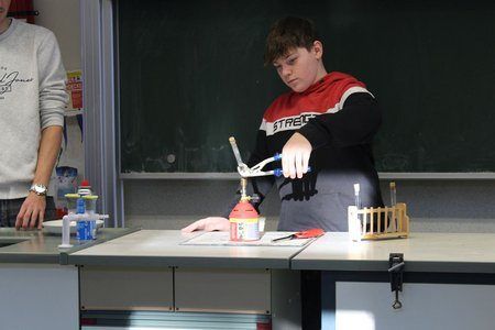 Ein Junge ist in einem Klassenzimmer und hält ein Werkzeug zum Reparieren einer Flamme. Er ist umgeben von Laborgeräten auf einem Tisch. Eine andere Person steht hinter ihm, in der Nähe eines Waschbeckens.