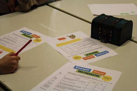 Tisch mit Escape-Room-Arbeitsblättern. Eine Person schreibt mit einem roten Stift auf ein Blatt Papier. Ein schwarzer Kasten mit Schlössern liegt auf dem Tisch.