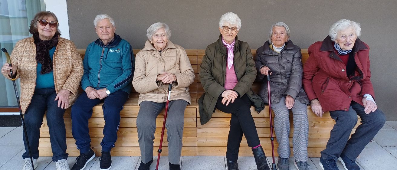 Eine Gruppe älterer Menschen sitzt draußen auf einer Holzbank. Sie tragen alle eine Brille und halten Gehstöcke. Sie sind warm gekleidet in Jacken und Hosen. Sie scheinen für ein Foto zu posieren.