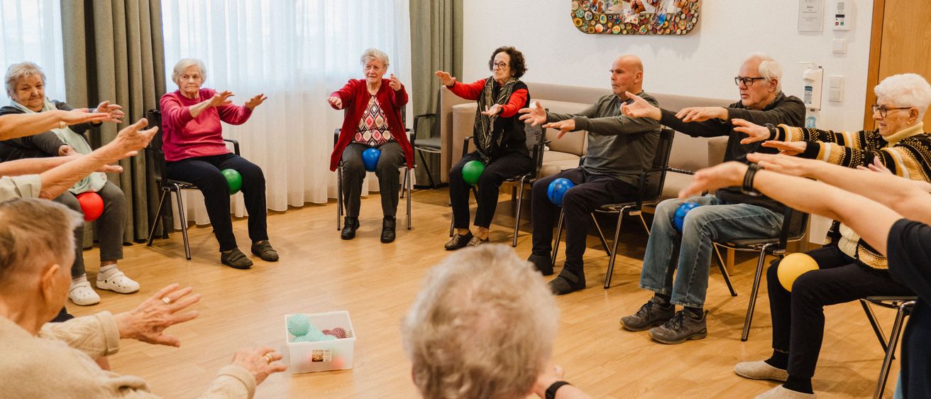 Eine Gruppe älterer Menschen nimmt an einer sitzenden Übungssitzung mit bunten Bällen teil, angeleitet von einem Trainer. Sie sitzen in Stühlen, die in einem Kreis angeordnet sind, mit einem dekorativen Wandbild im Hintergrund.