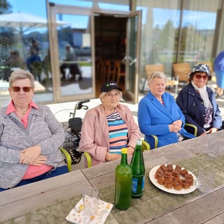 Vier ältere Frauen sitzen um einen Tisch herum außerhalb eines Gebäudes, mit Snacks und Getränken. Sie tragen Sonnenbrillen und Jacken. Hinter ihnen führt eine Glastür in einen Raum mit Stühlen und einem Tisch. Bäume und ein Auto sind draußen sichtbar.