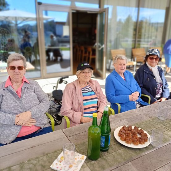 Vier ältere Frauen sitzen um einen Tisch herum außerhalb eines Gebäudes, mit Snacks und Getränken. Sie tragen Sonnenbrillen und Jacken. Hinter ihnen führt eine Glastür in einen Raum mit Stühlen und einem Tisch. Bäume und ein Auto sind draußen sichtbar.