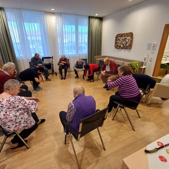 Eine Gruppe älterer Menschen sitzt in einem Kreis in einem Raum mit Fenstern und Holzböden. Einige schauen nach unten, während andere mit Aktivitäten beschäftigt sind.