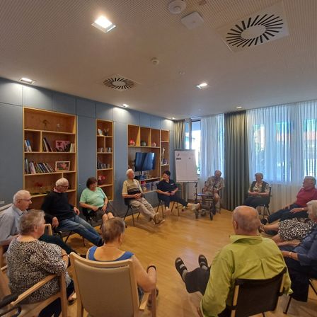Eine Gruppe älterer Erwachsener sitzt in einem Kreis in einem Raum mit Bücherregalen, einer Tafel und Fenstern mit Vorhängen. Einige sitzen in Rollstühlen.