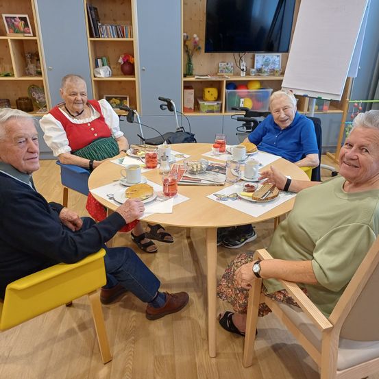 Vier ältere Personen sitzen in einem Wohnzimmer um einen Tisch herum, lächeln und genießen ihr Essen. Auf dem Tisch befinden sich Teller, Tassen und verschiedene Gegenstände.