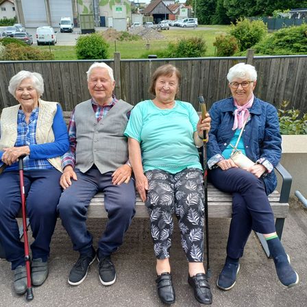Vier ältere Menschen sitzen auf einer Bank im Freien. Sie lächeln und posieren für ein Foto. Hinter ihnen steht ein Holzzaun. In der Ferne sind Autos geparkt. Bäume und ein Rasenfeld sind sichtbar.