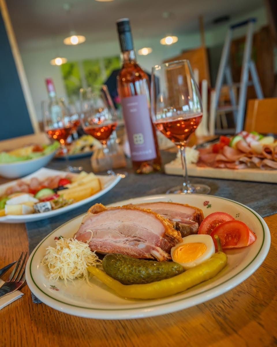 A wooden table is set with various dishes. A plate has ham, eggs, tomatoes, and pickles. A wine bottle and wine glasses are also on the table. Other plates contain assorted foods.