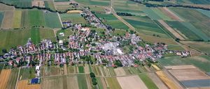 Luftaufnahme eines ländlichen Dorfes, umgeben von weitläufigen grünen Feldern, mit einer gewundenen Straße, die durch die Gemeinde führt.