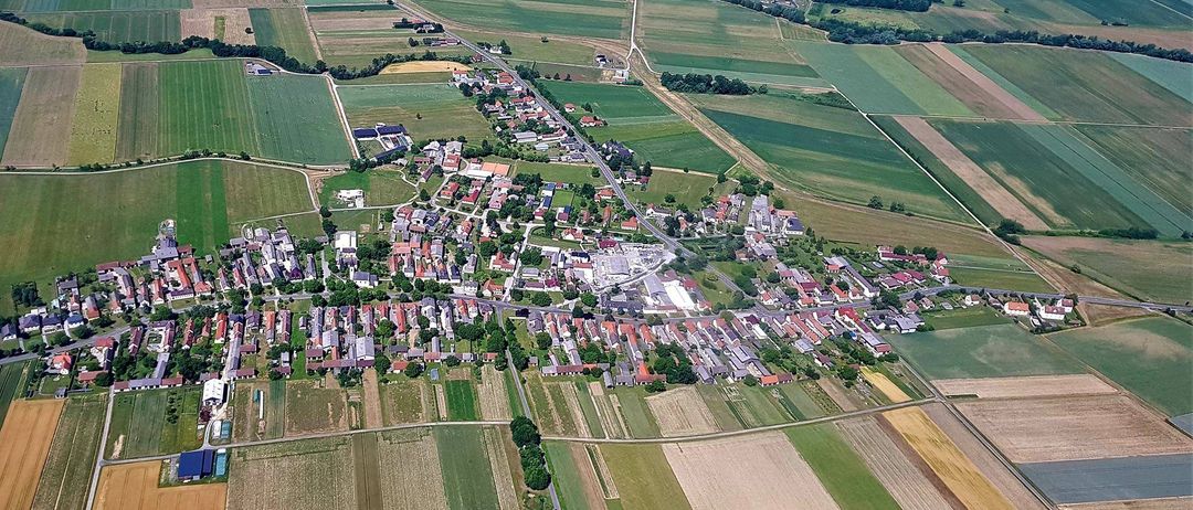 Luftaufnahme eines ländlichen Dorfes, umgeben von weitläufigen grünen Feldern, mit einer gewundenen Straße, die durch die Gemeinde führt.