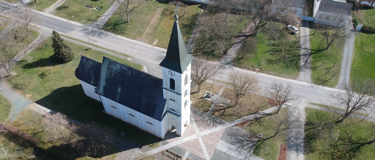 Luftaufnahme einer Kirche mit einem Turm und einem Glockenturm, umgeben von einem Kiesweg und grünem Rasen.