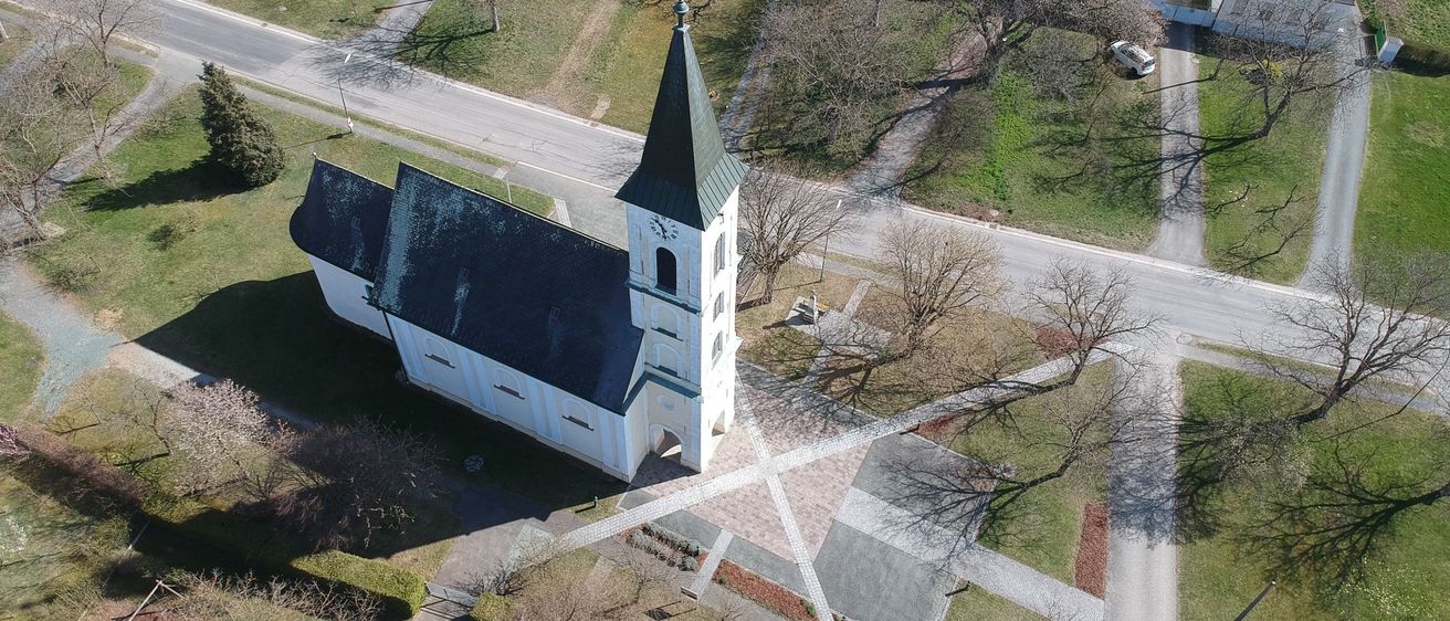 Luftaufnahme einer Kirche mit einem Turm und einem Glockenturm, umgeben von einem Kiesweg und grünem Rasen.