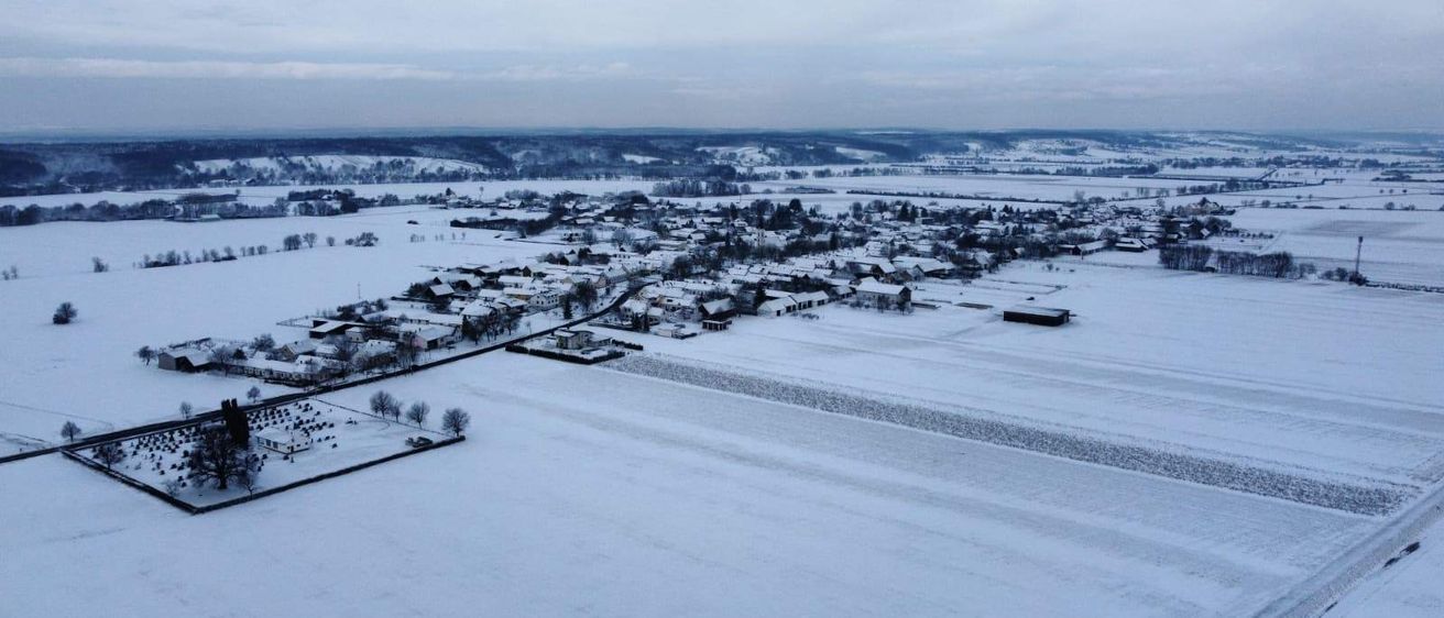 Luftaufnahme einer verschneiten Landschaft mit einem schneebedeckten Dorf und einem weitläufigen verschneiten Feld im Vordergrund.
