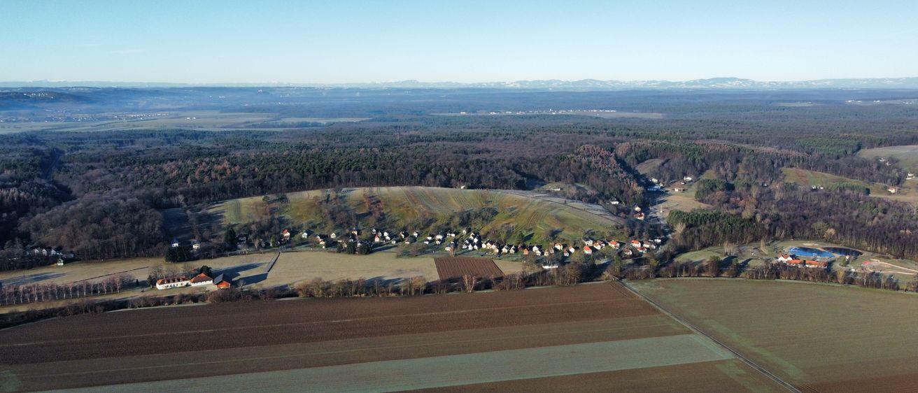 Luftaufnahme einer ländlichen Landschaft mit Häusern auf einem Hügel, umgeben von Feldern und Wäldern, unter einem klaren blauen Himmel.