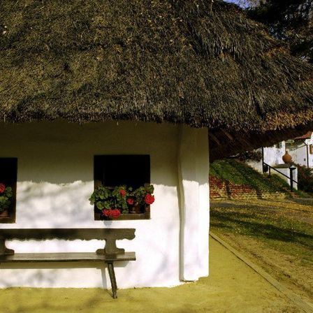 Ein kleines, weißes Häuschen mit einem strohgedeckten Dach, das zwei Fenster mit roten Blumen schmückt. Vor dem Häuschen steht eine Holzbank.