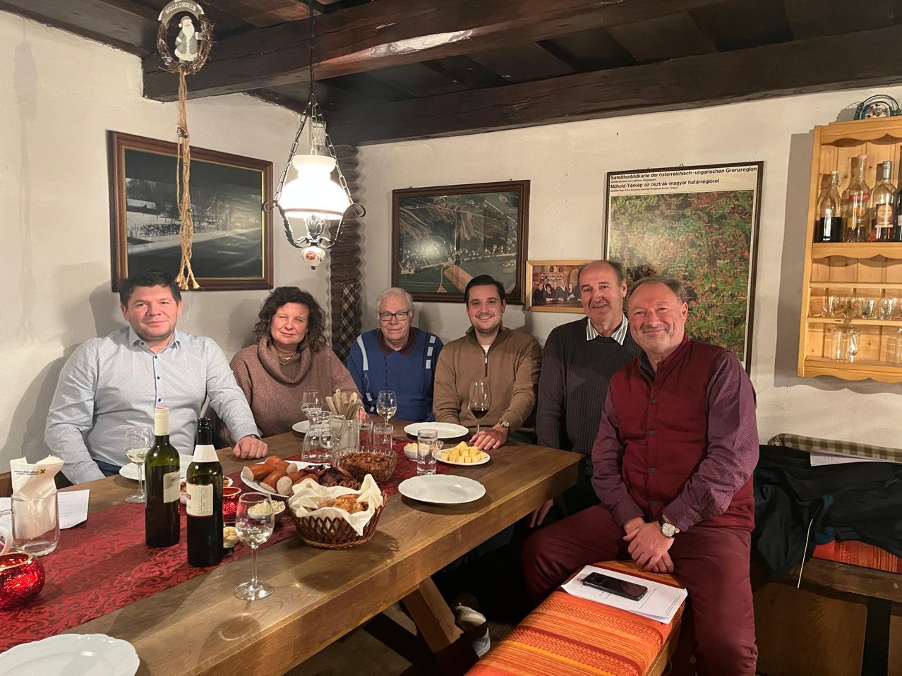 Sechs Menschen sitzen um einen Holztisch mit verschiedenen Speisen. Es gibt Teller, einen Korb und Weingläser. Sie lächeln und scheinen für ein Foto zu posieren. Der Raum hat Holzbalken, Gemälde und eine Lampe.