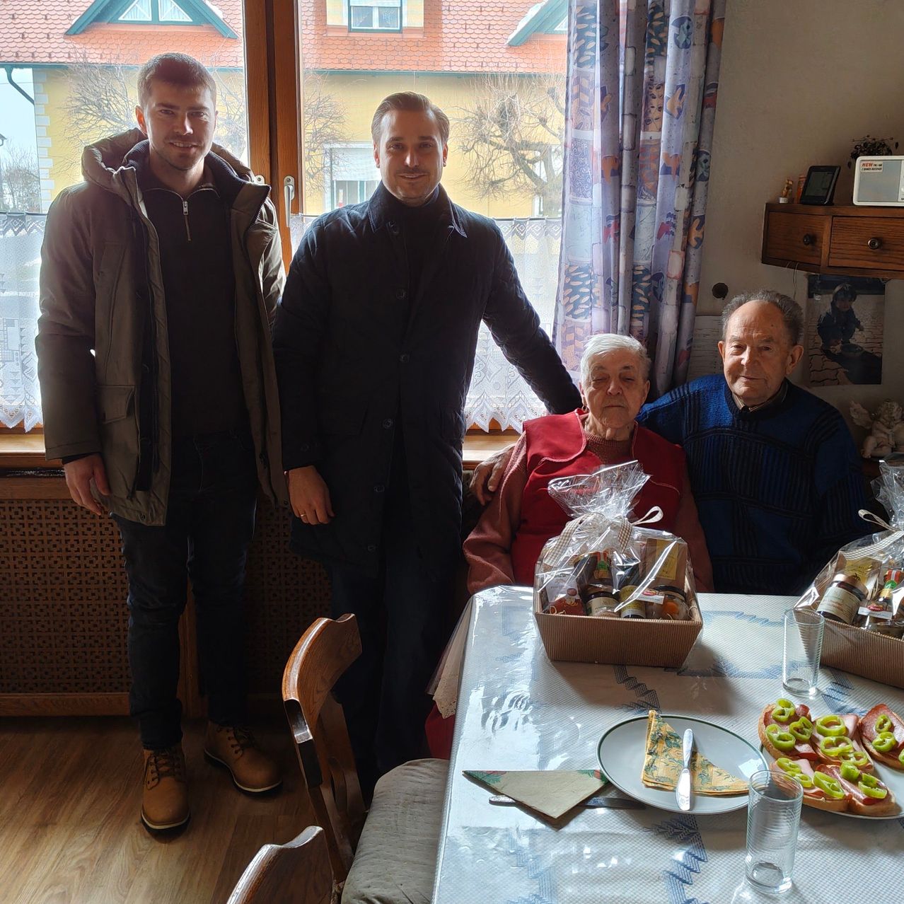 Drei Männer stehen in einem Raum, zwei vor einem Fenster, der dritte neben einer älteren Frau an einem Tisch mit Essen.