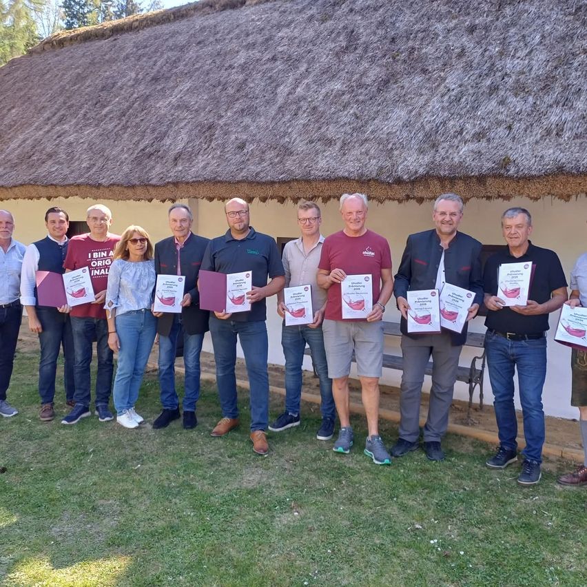 Eine Gruppe von Menschen steht vor einem Gebäude mit Strohdach und hält Urkunden. Sie lächeln und posieren für ein Foto.