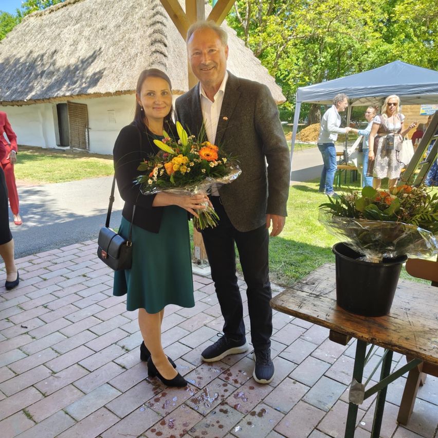 Ein Mann und eine Frau stehen auf einem Ziegelboden und lächeln, während sie einen Blumenstrauß halten. Dahinter steht ein Haus mit Strohdach und ein Zelt.