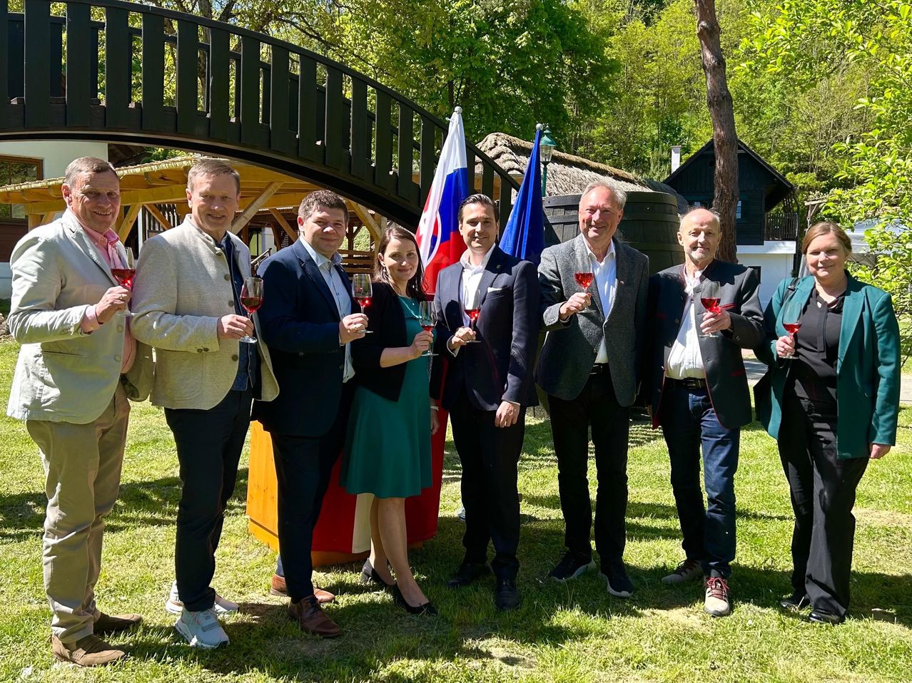 Gruppe von Menschen in formeller Kleidung, die in einem Grasbereich stehen und Weingläser halten. Hinter ihnen sind eine Brücke und Fahnen.
