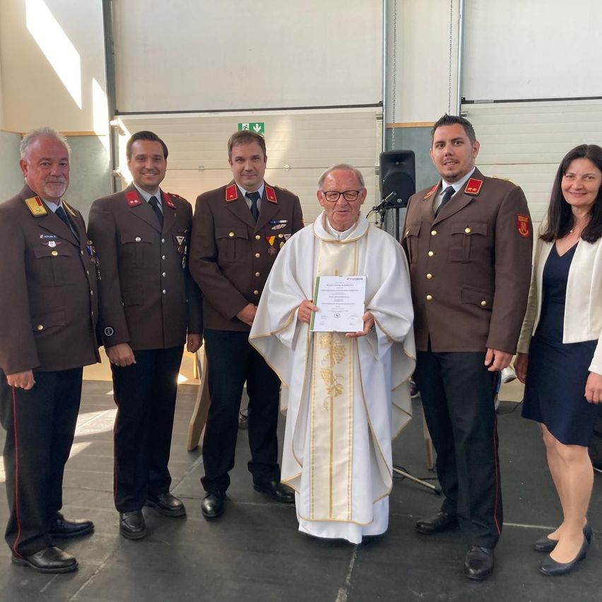 Eine Gruppe von Menschen, darunter ein Priester und Militäroffiziere, steht in einem Saal zusammen und lächelt für ein Foto. Der Priester hält eine Urkunde.