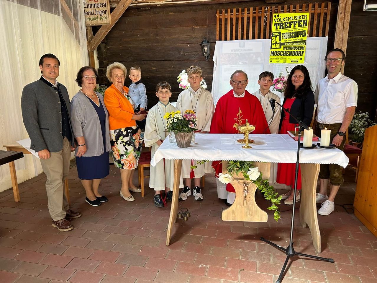 A group of people stands together in front of a church altar, with two boys in white robes and two candles. A priest is standing behind the altar, holding a microphone.
