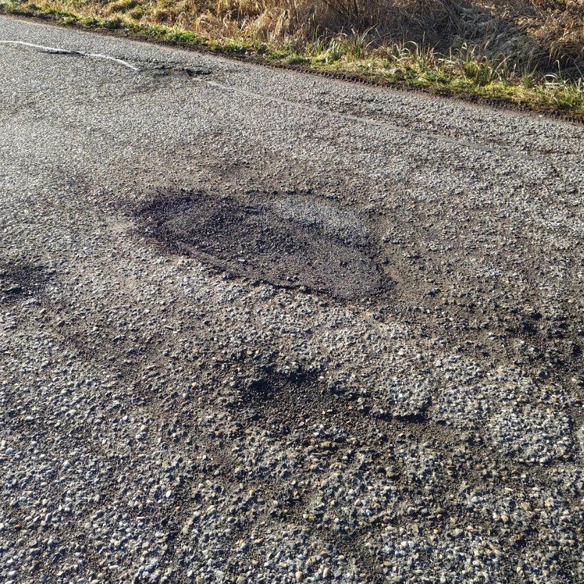 A gravel road with a noticeable pothole near the edge, surrounded by sparse grass and dry vegetation.