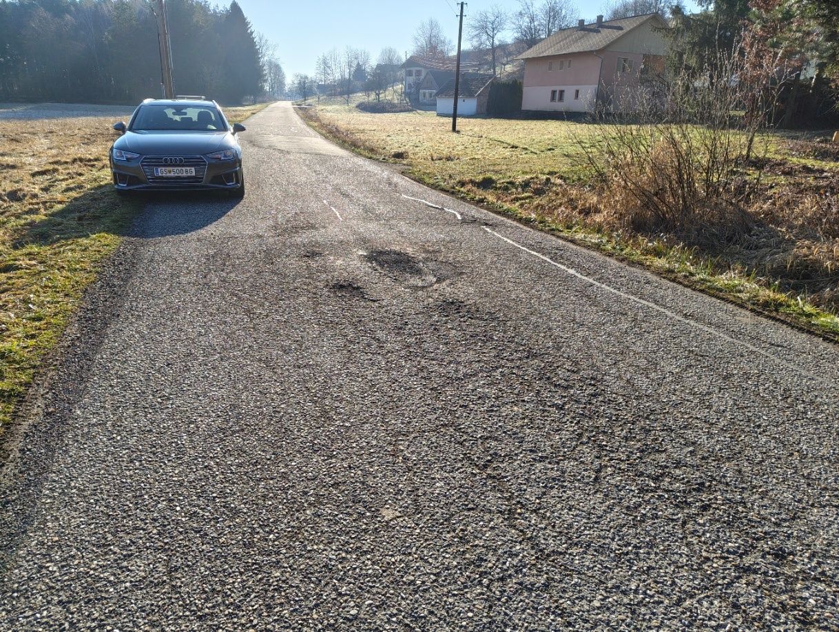 A dark colored Audi car with the license plate GS 500 B6 parked on the side of a road. The road is made of gravel with potholes and surrounded by grass.