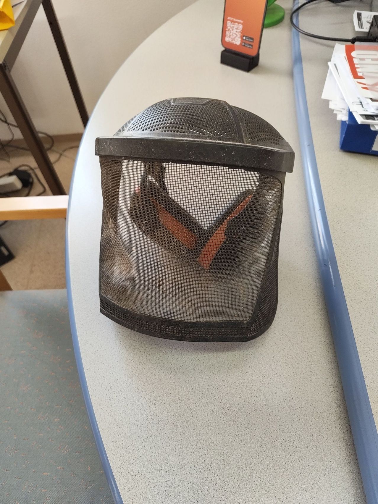 Ein Schutzhelm mit schwarzem Netz und orangefarbenen Ohrenschützern steht auf einem grauen Tisch. Im Hintergrund befinden sich Papiere und ein blaues Objekt.