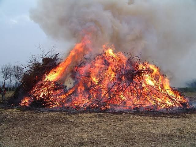 Ein großes Feuer brennt auf einem Feld mit viel Rauch. Einige Leute stehen in der Nähe.