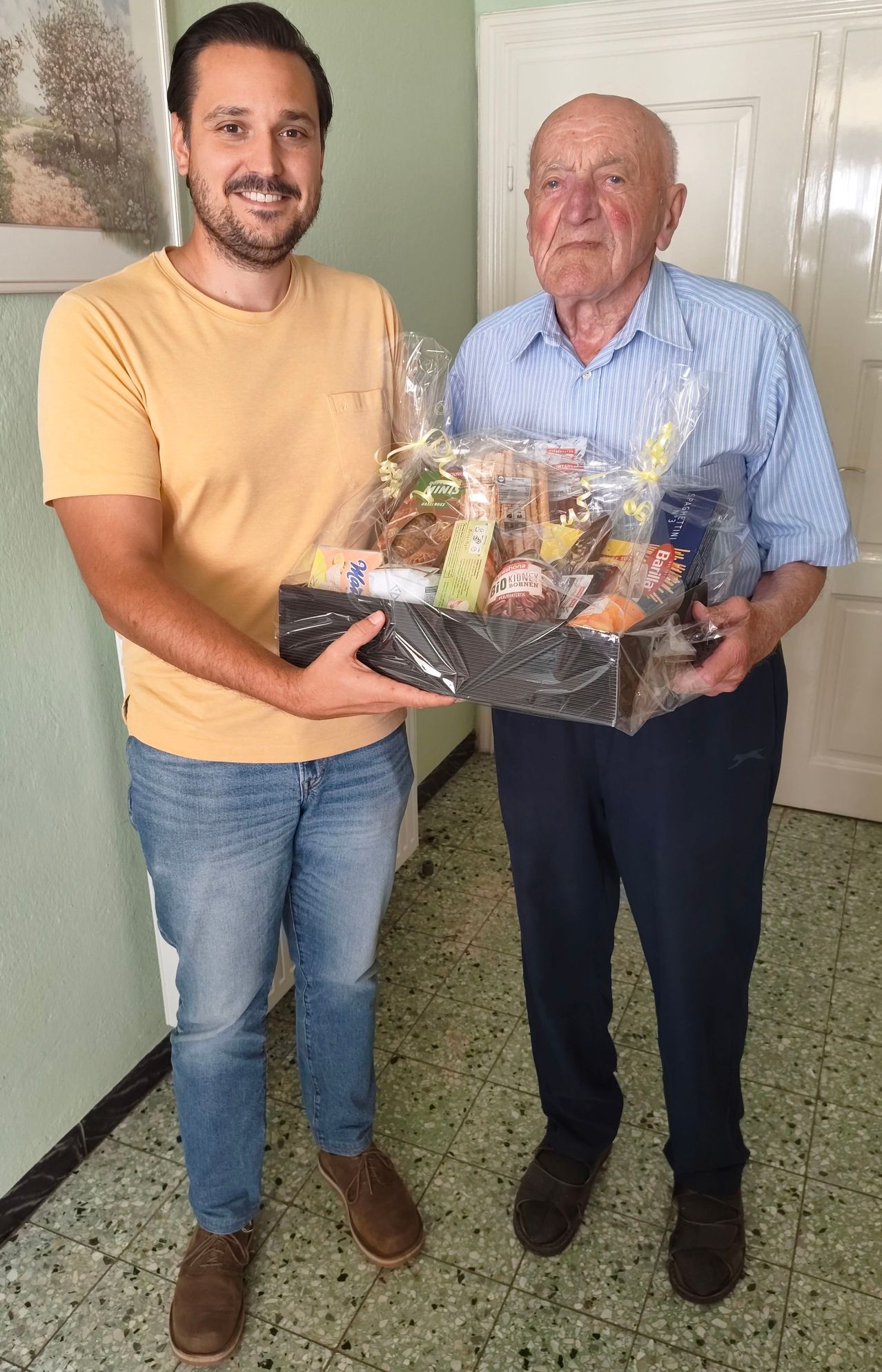 Zwei Männer halten einen Geschenkkorb mit verschiedenen Snacks in einem Flur.