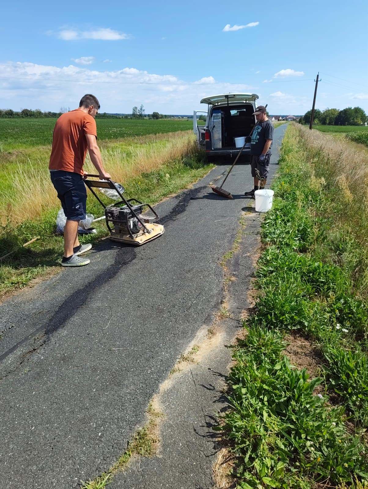 Zwei Männer arbeiten an einer Straße, einer benutzt eine Maschine und der andere hält einen Eimer. Die Straße ist von Gras umgeben und ein Van ist in der Nähe geparkt.