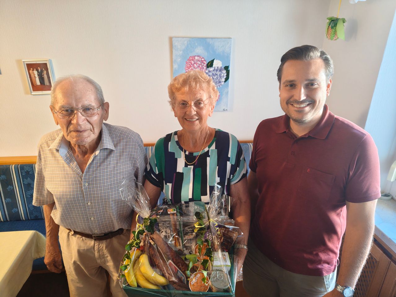 Drei Menschen, zwei ältere Erwachsene und ein junger Mann, lächeln und posieren für ein Foto, während sie einen Geschenkkorb mit Obst und Snacks halten.