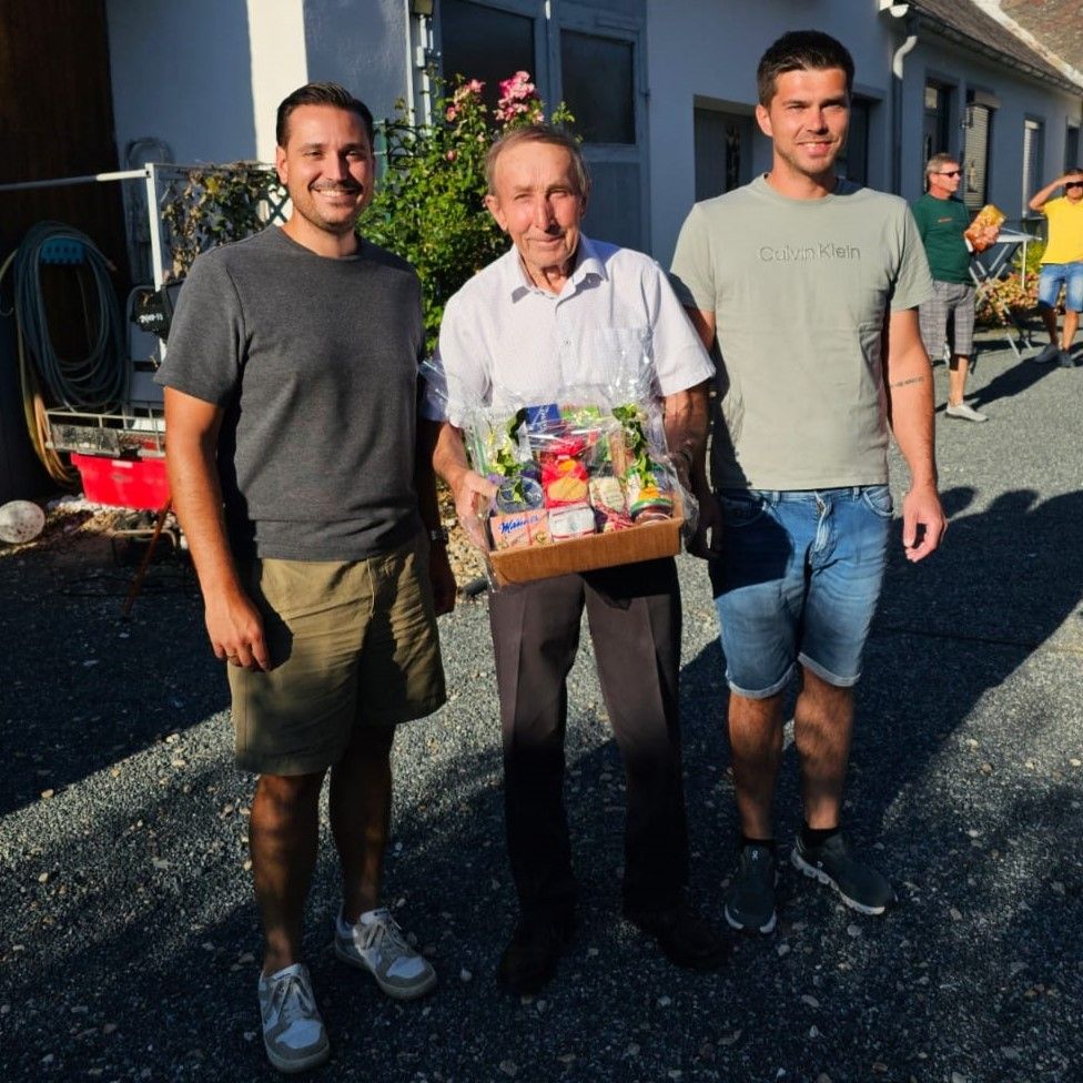 Three men stand outside, one holding a box of snacks. The man in the middle wears a white shirt, while the others wear gray and green shirts. Behind them is a house with a garden.