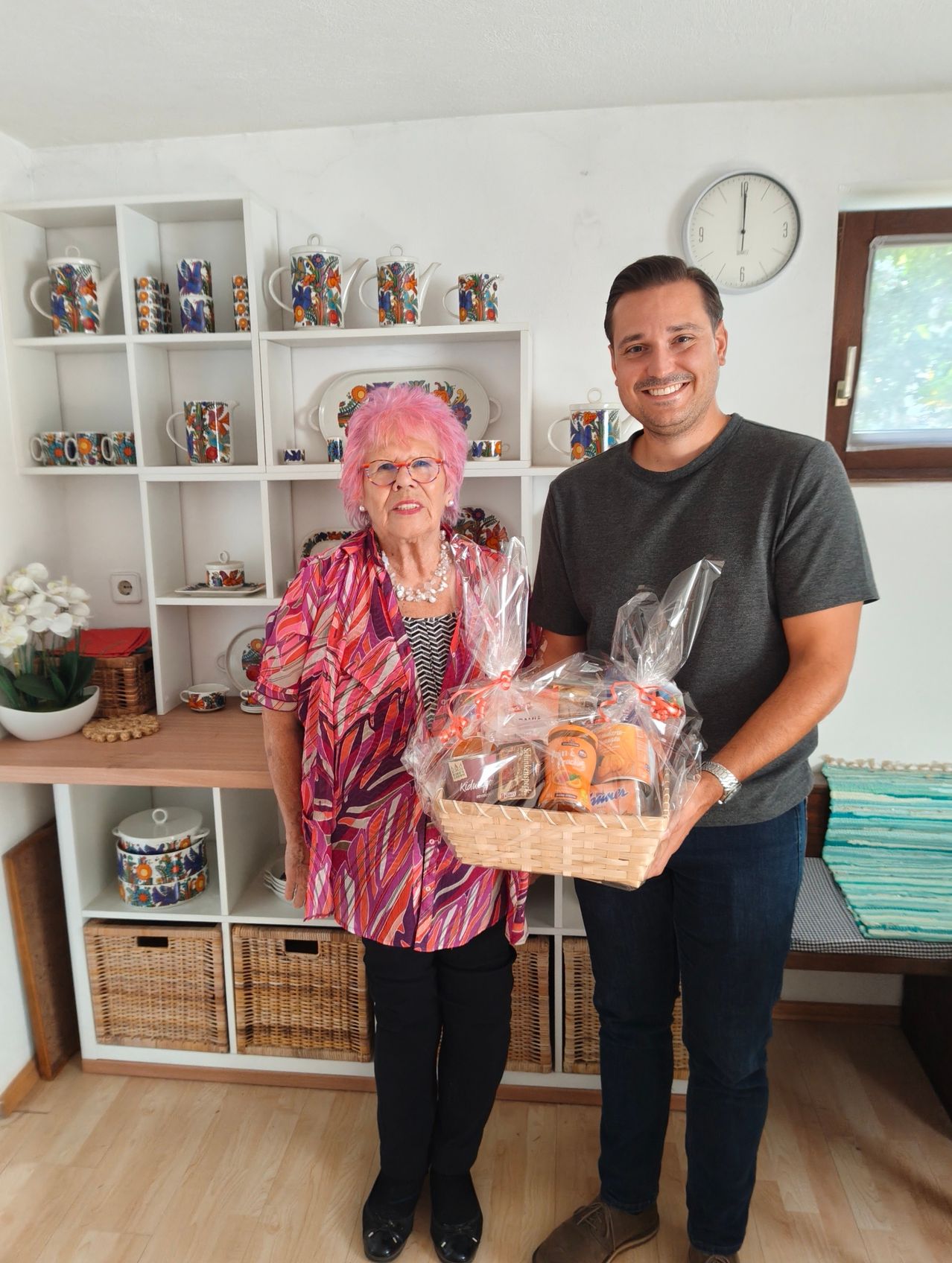 A man and an elderly woman are standing in a room, holding a basket of food. The woman has pink hair and is wearing a colorful blouse. The man is wearing a gray t-shirt and jeans. Behind them is a shelf with various items.