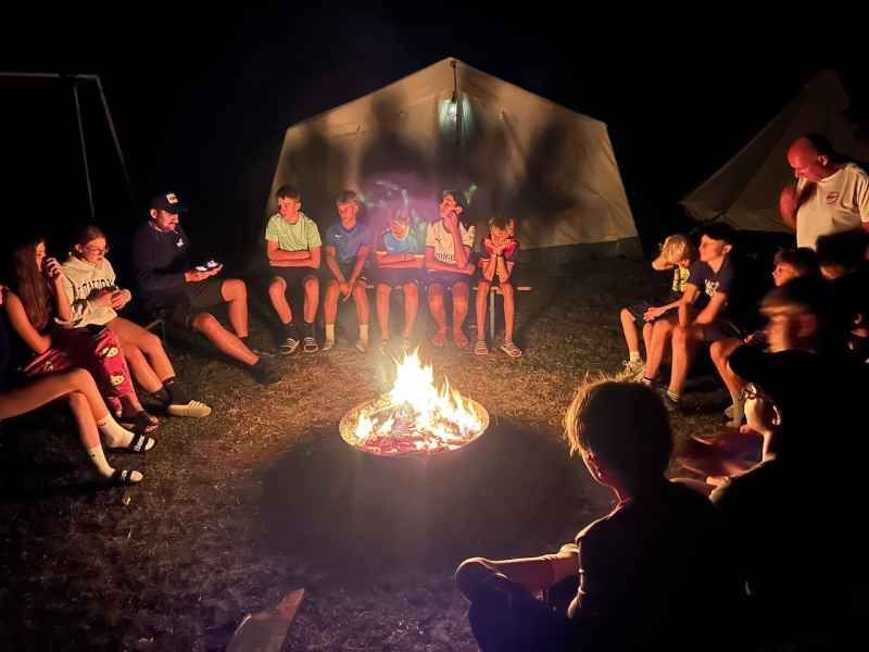 A group of people, including children, are gathered around a bonfire at night, sitting on the ground and on a bench, with tents in the background.