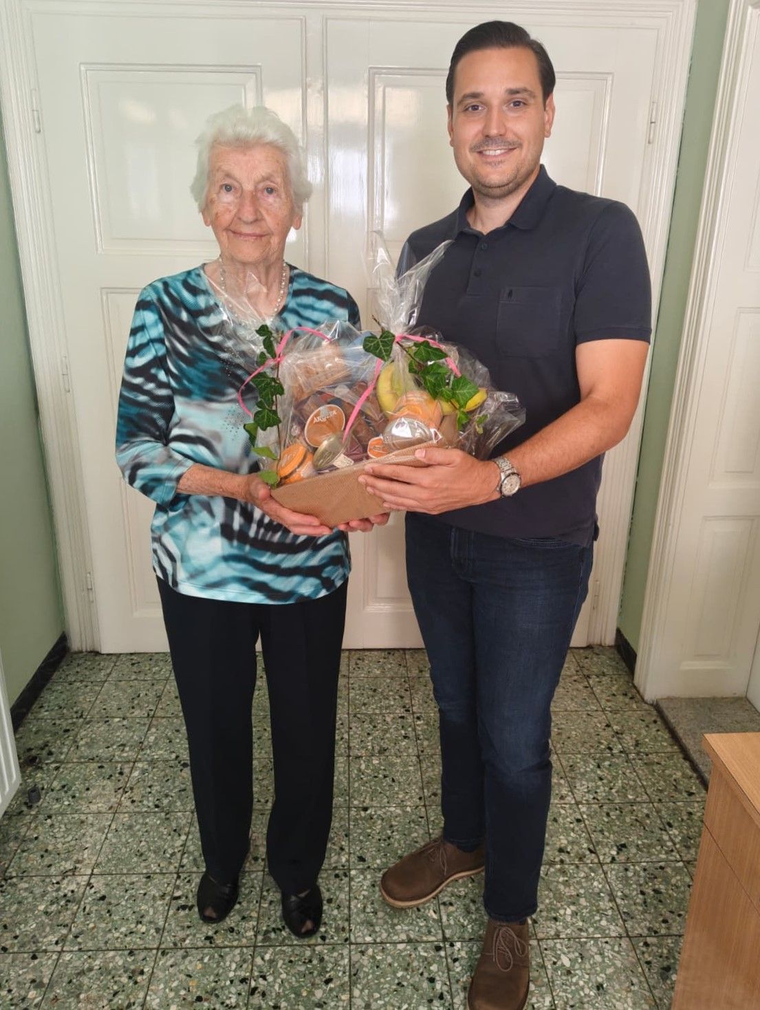 An elderly woman and a man are standing in a hallway, smiling. The man is holding a large gift basket filled with fruits and other items. They are both dressed casually.