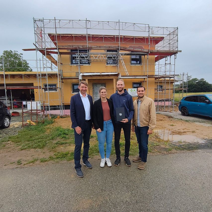 Four people stand in front of a construction site, smiling, with a woman holding a certificate. Behind them is a yellow building under construction.