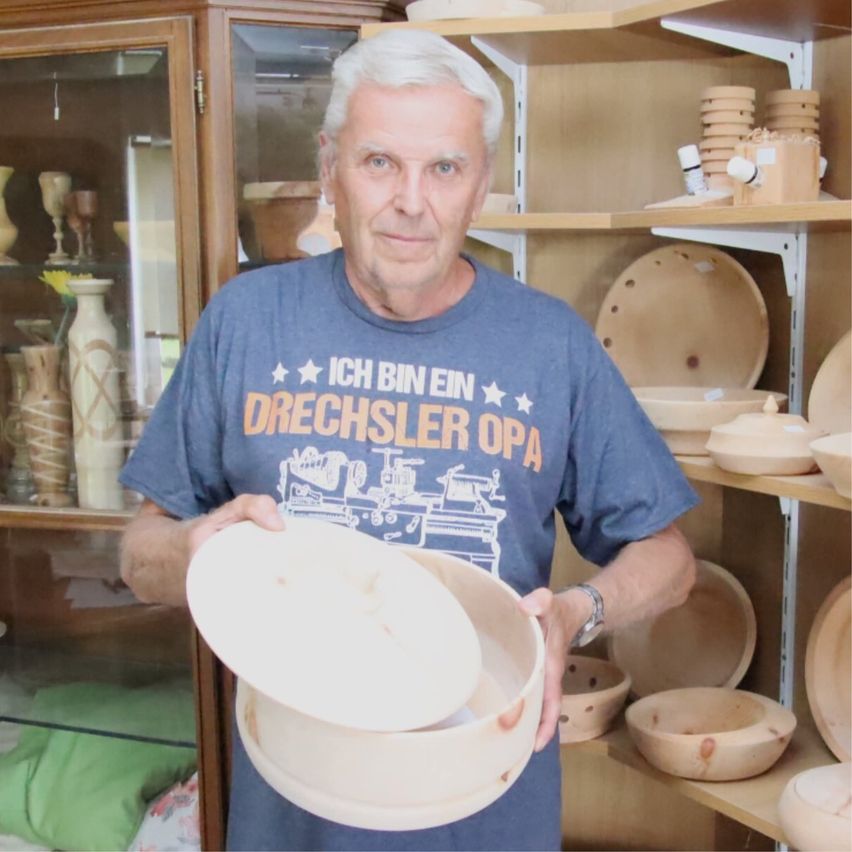 An older man in a blue shirt with a woodturning design is holding a wooden bowl in a room full of wooden bowls and vases.