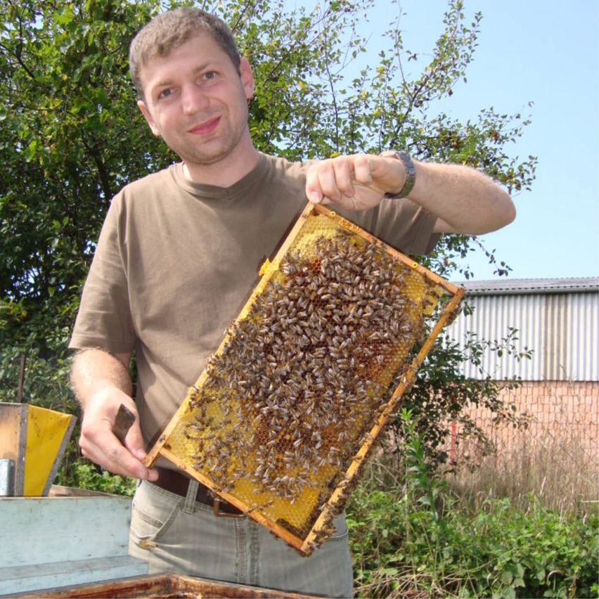 A man in a brown shirt and watch holds a honeycomb filled with bees outdoors, smiling for the camera.