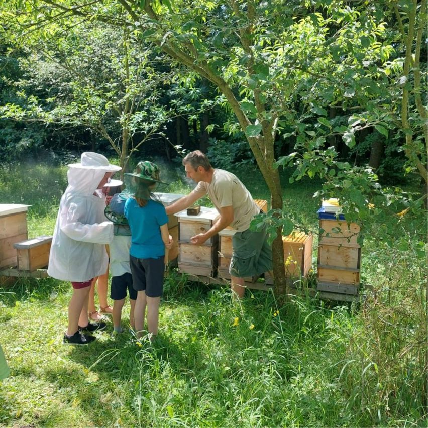 In a garden, a beekeeper in a protective suit helps a child in a hat. Others watch as they work near wooden bee boxes.
